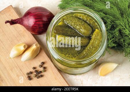 Delicious pickled cornichons in an open glass jar, dill, onion, garlic and pepper on a table. Whole green gherkins marinated with dill and mustard see Stock Photo
