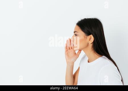 Side view young beautiful woman making announcement or telling secret holding her hand to her face on white studio background with copy space. Stock Photo