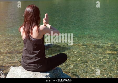 Young sporty woman practicing yoga in black sportswear near the river or lake with transparent water Stock Photo