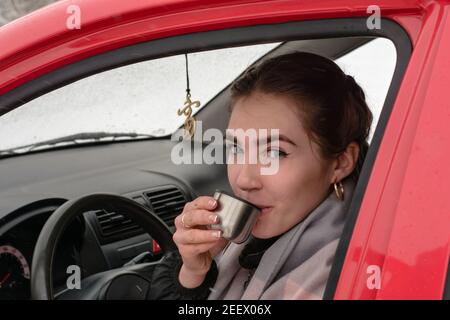 Young woman driving red car drinks hot coffee from thermos mug. Travel rest concept. Stock Photo