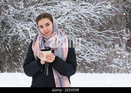 Fashioning beautiful woman in winter, scarf on head holds mug with hot tea in  hands. Branches of trees  hoarfrost and snow forest. Stock Photo