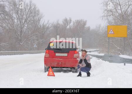 Car breakdown on track in  blizzard and bad weather. Woman driver with no experience sits and worries near triangular accident sign. Stock Photo