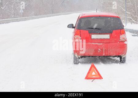 Broken car on snowy road. Red triangle warning sign for emergency stop. Snow and blizzard, winter driving hazard. Stock Photo