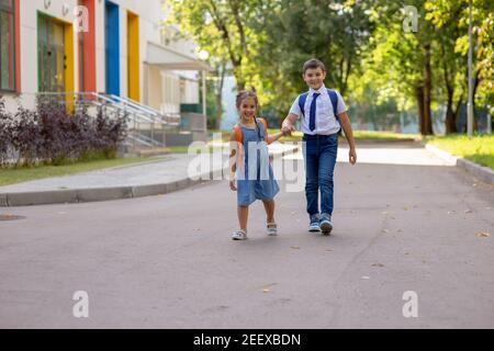 Cheerful schoolchildren, a girl and a boy in a white shirt with backpacks, go from school. Stock Photo