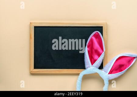 Blank blackboard and bunny ears costume headband on beige background. Easter concept. Top view, flat lay, mock up. Stock Photo