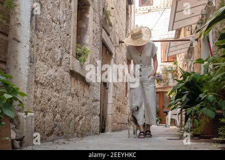 Rear view of beautiful blonde young female traveler wearing straw sun hat sightseeing and enjoying summer vacation in an old traditional costal town Stock Photo