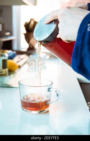 Close up man in home bathrobe pouring hot tea from glass teapot into cup on the kitchen table. Process brewing tea in the morning. A cup of freshly Stock Photo