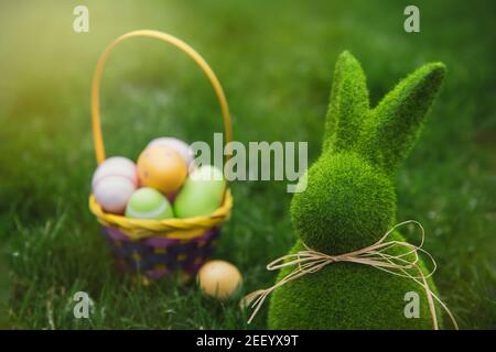 Close up Easter bunny rabbit statuette and basket with easter eggs on the green grass lawn background. Easter egg hunt in the garden. Selective focus Stock Photo