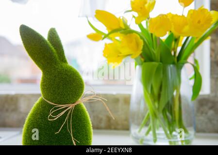 Cute green grass bunny rabbit and bouquet of fresh spring yellow tulips and daffodils flowers on the white kitchen table near the window. Happy Easter Stock Photo