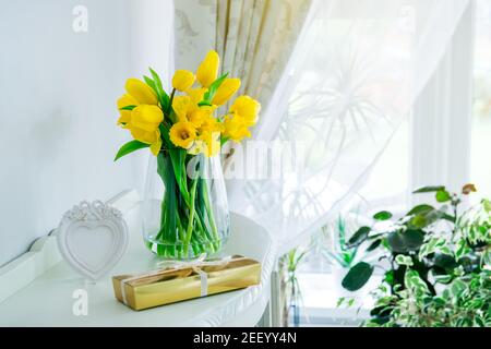 Bouquet of fresh yellow spring tulips and daffodils flowers, golden gift box and heart shaped frame on the chest drawers in light classic interior Stock Photo