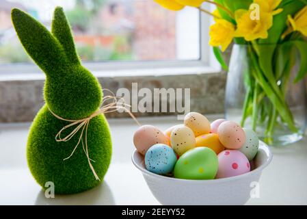 White bowl with colored easter eggs, bouquet of yellow tulips and daffodils flowers and green bunny rabbit on white kitchen table near window. Festive Stock Photo
