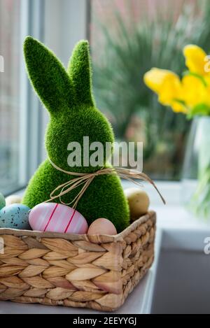 Close up Easter bunny rabbit in straw basket with colored eggs on the windowsill with fresh spring tulips and daffodils flowers bouquet on the Stock Photo