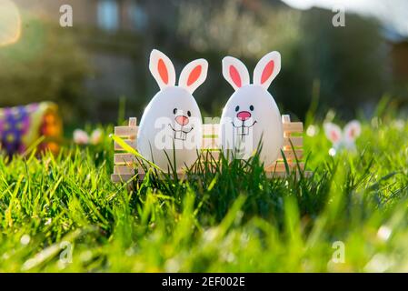 Creative Easter composition of Funny Easter eggs with bunny ears sitting on bench on green grass background with fallen basket. Sunny festive mood Stock Photo