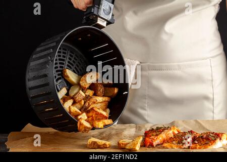 A caucasian woman chef is dumping fresh made fried potato chunks on a piece of baking paper which has three oven roasted marinated salmon fillet with Stock Photo