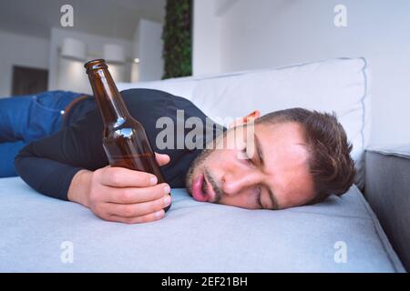 Drunk young man with open mouth and bottle of beer sleeping on the sofa at home. Stock Photo