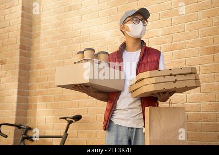 Male courier wearing face protective mask holding various packages with food, pizza boxes and four coffee cups while standing against brick wall. Delivery services during Covid 19 outbreak Stock Photo