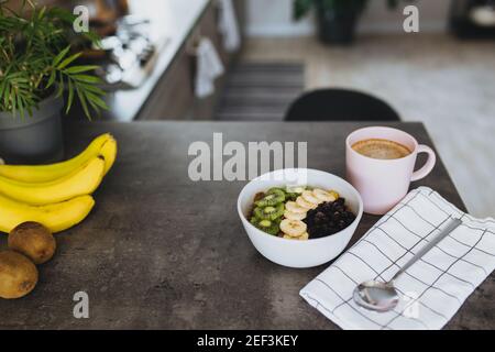 Pink coffee cup, bowl with chopped tropical fruits kiwi and banana, blueberries, spoon on bar counter in stylish loft kitchen. Blurred background. Hig Stock Photo