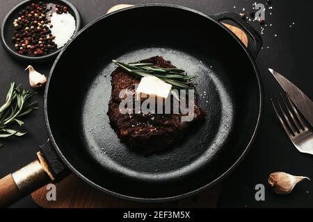 Cooked ribeye steak on cast-iron pan and board with pepper, rosemary, salt, garlic, knife, fork, butter on black background Stock Photo