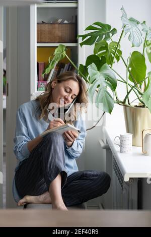 Smiling woman talking on cellphone writes in a notebook, sitting next to the window at home. Stock Photo