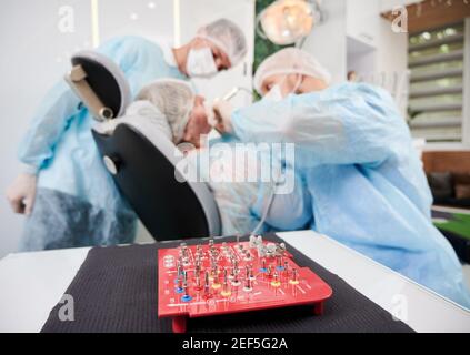 Close up of dental implant surgical kit with doctors and patient on blurred background. Dentist using dental implant machine while performing implantology procedure in dental office. Stock Photo