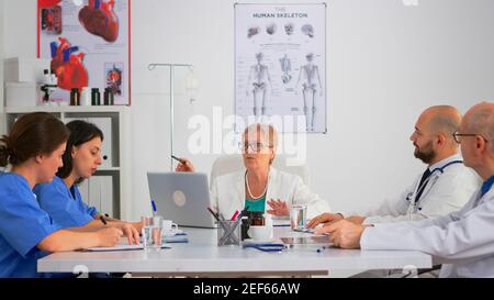 Elderly specialist woman doctor sitting on conference desk talking with team during medical meeting about medical procedures while coworkers taking notes. Profesional team having brainstorming. Stock Photo