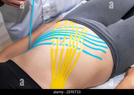 Male physiotherapist applying blue adhesive elastic therapeutic tape on female abdominal muscle of young adult slim sporty woman lying on couch doctor Stock Photo