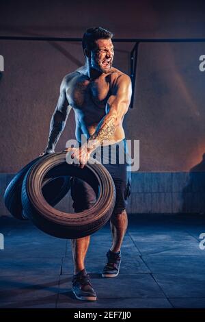 Bodybuilding training, bearded strong sportsman with muscular body lifting heavy wheel in gym. Stock Photo