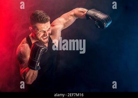 Bearded tattooed sportsman muay thai boxer in black undershirt and boxing gloves fighting on dark background with smoke. Sport concept. Stock Photo