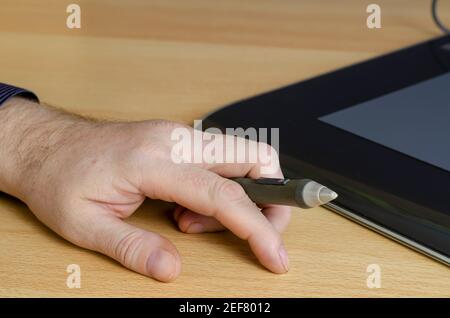 A man's hand with a special pen for a graphics tablet in close-up. A professional device for designers lies in the background. Side view from an angle Stock Photo