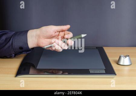 A man's hand with a special pen on top of the screen of a graphics tablet. A professional device for designers lying on a table. Side view from an ang Stock Photo