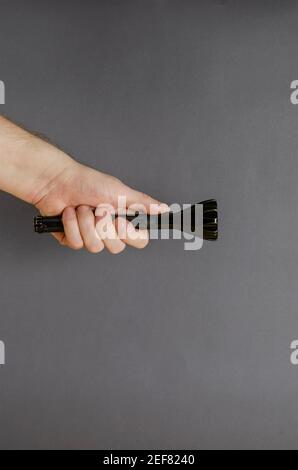 A hand holds a black modern LED flashlight on a gray background. Male hand and a metal flashlight. Side view. Selective focus. Stock Photo