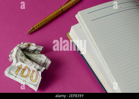 Open diary, gold pen and crumpled one hundred US dollar bill on pink background. Top view at an angle. Selective focus Stock Photo