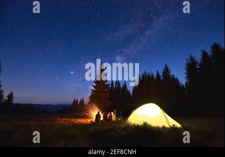 Evening camping near fire, spruce forest and mountains on background. Group of friends having a rest near bright bonfire. People sitting near tourist tent under night sky full of stars and Milky way. Stock Photo