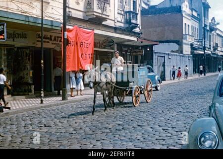 Milk being delivered by a cart being pulled by a donkey on a cobbled street in Petrópolis (also known as The Imperial City), Brazil 1961. It was common for many fresh goods were delivered door to door by carts pulled by animals or by motorised vehicles. On the side of the cart is the word ‘Leite’ – Portuguese for milk. The city of Petrópolis is located 68 km (42 ml) north east of Rio de Janeiro. It is the largest and most populous city in the Fluminense Mountain Region. This image is from an old amateur 35mm colour transparency. Stock Photo