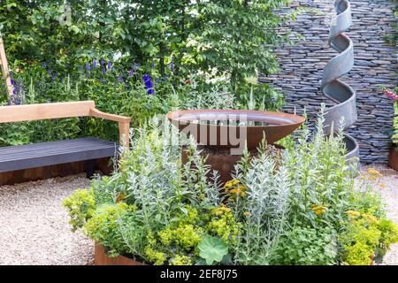 Small English courtyard garden with spiral sculpture - corten steel water feature - hedges - gravel path with garden bench flower borders England UK Stock Photo