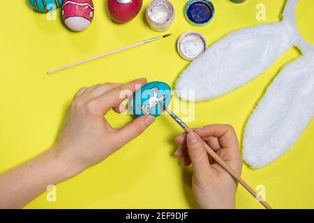 A woman paints an Easter egg with gouache paints. Painting eggs and preparing for Easter. An image of a rabbit on an egg on a yellow background. Stock Photo
