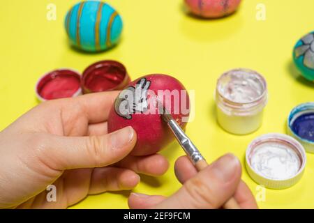 A woman paints a chicken egg with gouache paints with a brush on a yellow background. Easter photo, preparation for the holiday. Stock Photo