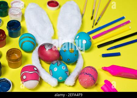 Easter concept: bunny ears, painted eggs, paints, felt-tip pens and markers on a yellow background. Preparing for the Easter holiday Stock Photo
