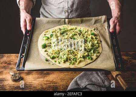 Man holding baking dish with uncooked pizza with broccoli, pesto sauce, spices and cheese Stock Photo