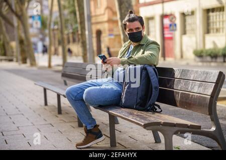 Young man wearing a mask and sitting on the bench with his backpack Stock Photo