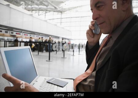 Side profile of a businessman talking on a mobile phone and using a laptop at an airport lounge Stock Photo