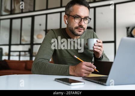 Handsome confident young businessman working in a modern office space, using laptop computer, sitting at the desk, taking notes Stock Photo