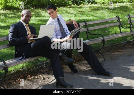 Two businessmen sitting on a bench in a park and looking at a laptop Stock Photo