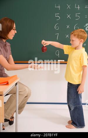 Side profile of a schoolboy giving an apple to his female teacher in a classroom Stock Photo