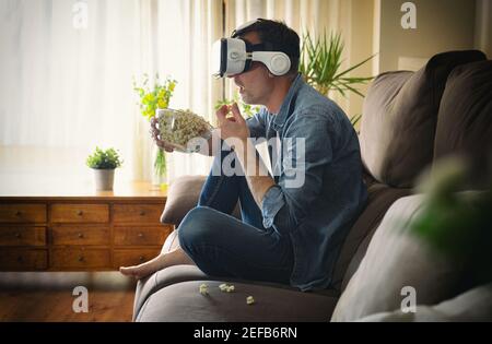 Astonished man watching a movie in virtual reality glasses and eating popcorn sitting on the sofa at home Stock Photo