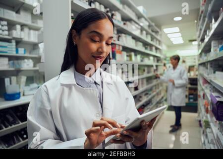 Smiling young female worker in pharmacy wearing labcoat checking inventory using digital tablet Stock Photo