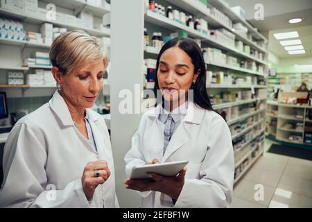 Happy young female pharmacist discussing work with senior colleague using digital tablet in chemist Stock Photo