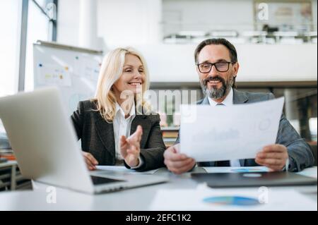 Joyful successful coworkers doing some paperwork in modern office, looking at the financial graphs. Happy mature stylish partners sitting at the desk, working on new business plan Stock Photo