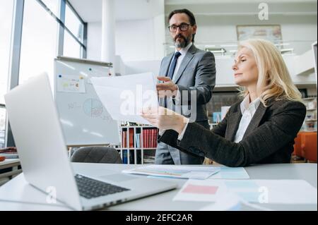 Serious mature male and female is learning financial graphs, doing paperwork at the office. Beautiful blonde hair woman and attractive man working together on important business project Stock Photo