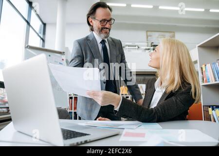 Friendly middle aged colleagues working in modern office, discussing new business plan. Pretty woman in formal wear ask for advice about some documents or financial graphs from smart male colleague Stock Photo
